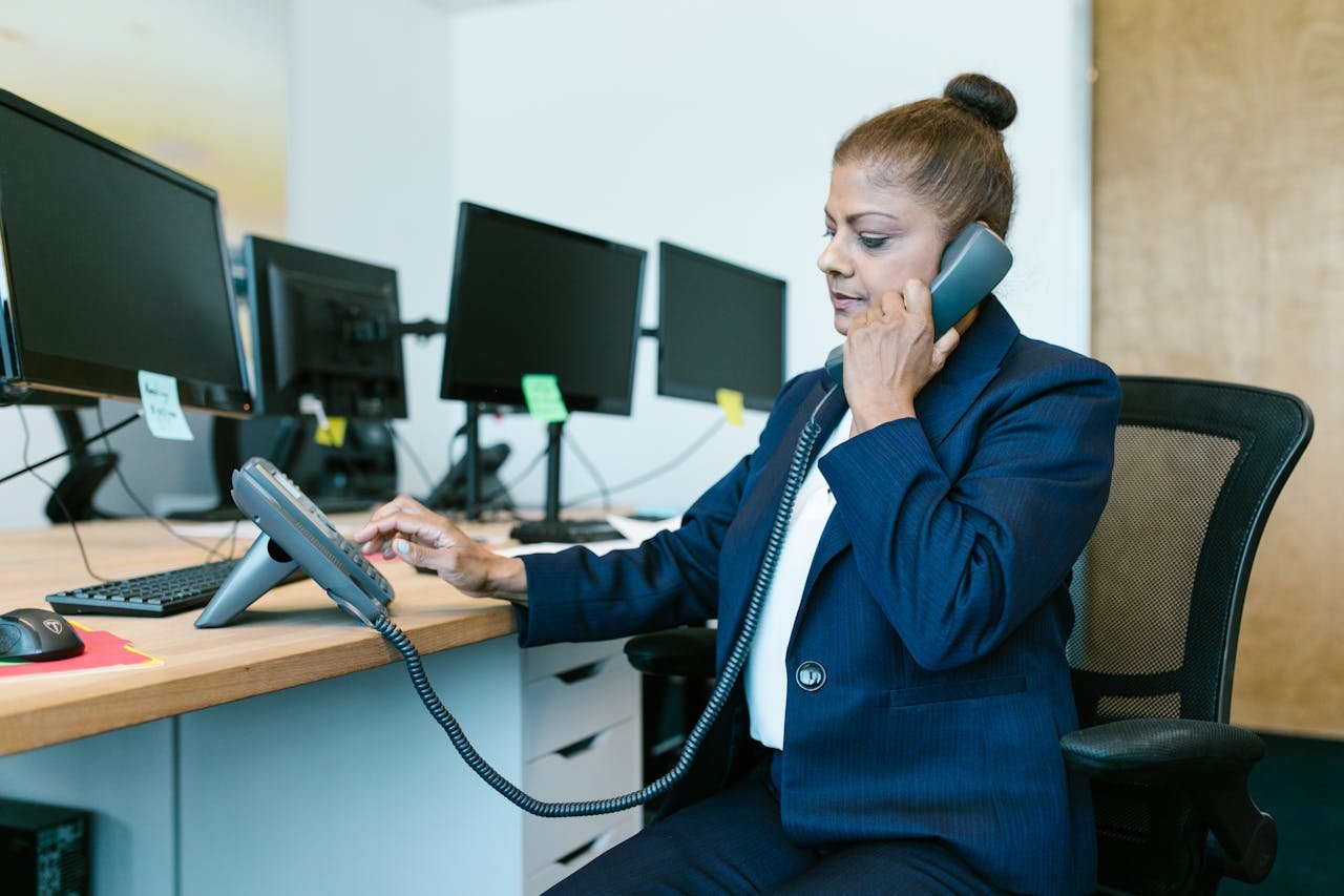 Businesswoman multitasking in office, using phone and computer, wearing formal attire.